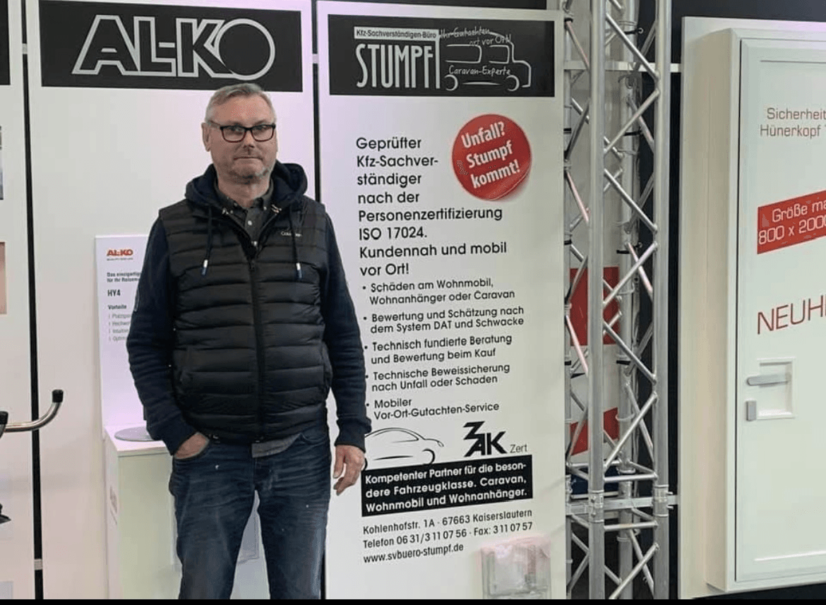 Rainer Stumpf steht vor einem Messestand seines Kfz-Sachverständigenbüros „STUMPF“ auf einer Fachmesse. Er trägt eine dunkle Daunenweste mit Kapuze, Jeans und eine Brille. Hinter ihm ist ein Roll-up mit den angebotenen Leistungen zu sehen: mobile Gutachten und Bewertungen speziell für Wohnmobile, Wohnanhänger und Caravans. Der Slogan „Unfall? Stumpf kommt!“ sticht in einem roten Kreis hervor. Auf der linken Seite ist das AL-KO-Logo sichtbar, rechts ein weißer Ausstellungsstand mit roter Schrift.