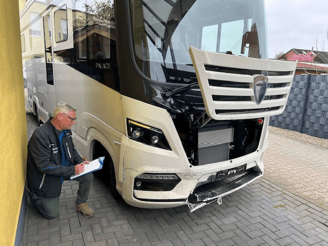 Rainer Stumpf kniet neben einem beschädigten weißen Luxus-Wohnmobil der Marke Morelo mit der Modellbezeichnung „PALACE“ und dokumentiert den Frontschaden auf einem Klemmbrett. Die Frontverkleidung des Fahrzeugs ist stark beschädigt und teilweise demontiert, der Kühler ist sichtbar. Rainer trägt eine dunkelgraue Jacke mit Aufdruck „SV Büro Stumpf“ und grüne Hose. Die Szene findet auf einem gepflasterten Hof statt, neben einem gelben Haus.