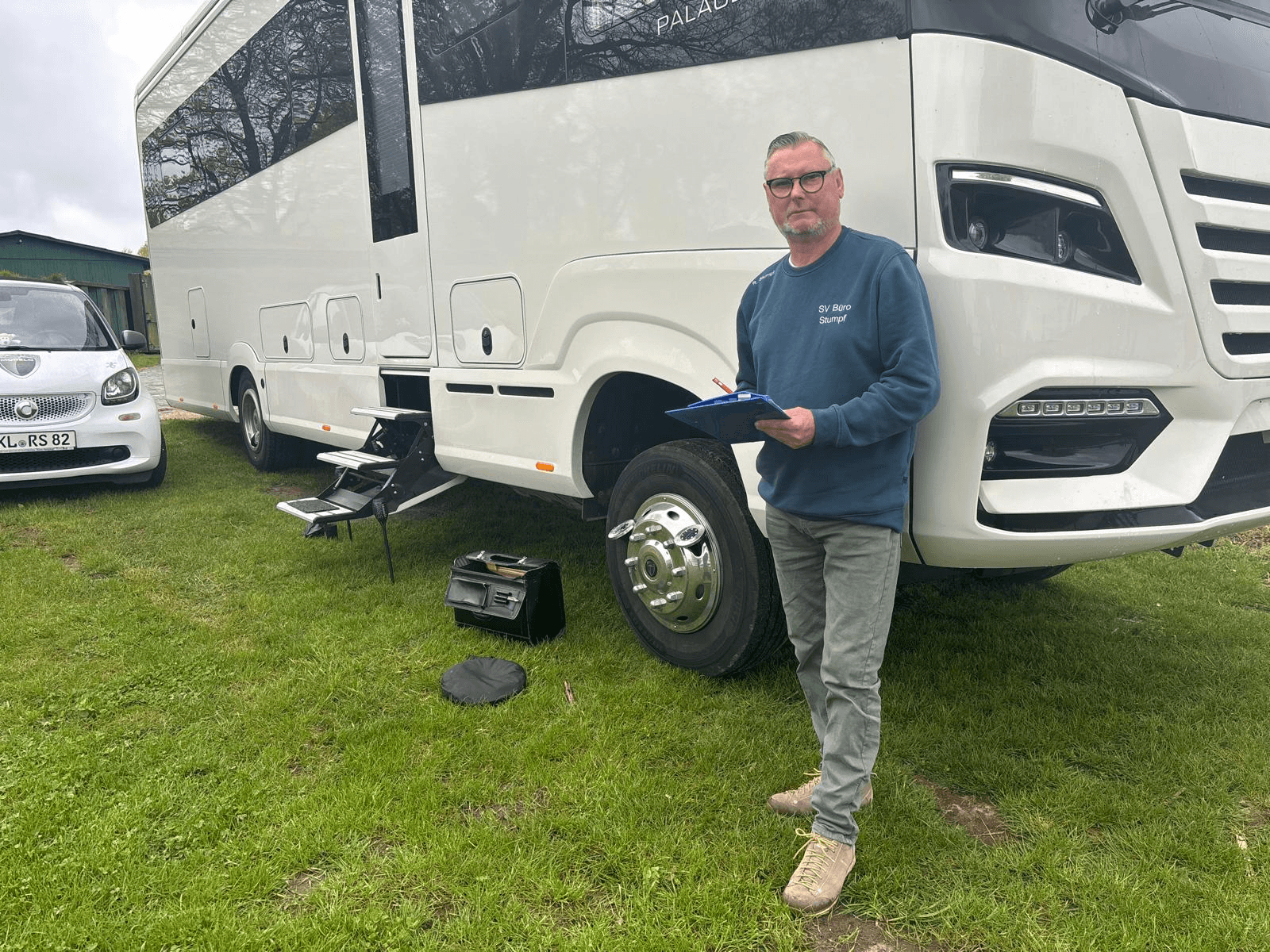 Rainer Stumpf, Kfz-Sachverständiger, steht mit Klemmbrett und Stift neben einem weißen Luxus-Wohnmobil auf einer Wiese. Er trägt eine dunkelgrüne Hose und ein Sweatshirt mit der Aufschrift „SV Büro Stumpf“. Im Vordergrund liegen eine schwarze Werkzeugtasche und ein Sitzkissen. Links im Bild parkt ein weißer Smart mit dem Kennzeichen KL-RS 82. Die Szene wirkt wie eine Schadensaufnahme oder Begutachtung vor Ort.
