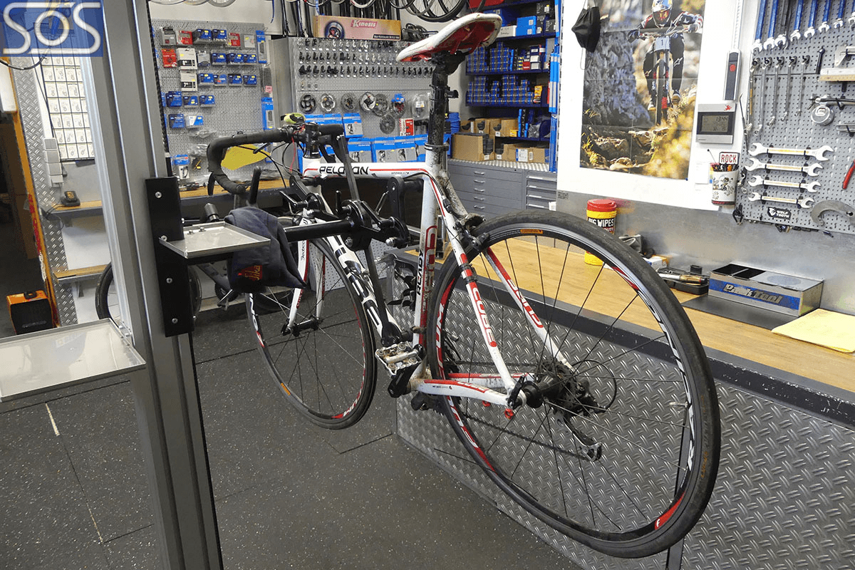 Ein weiß-rotes Rennrad der Marke Cube ist in einer Fahrradwerkstatt in einem Prüfstand eingespannt. Im Hintergrund befinden sich zahlreiche Werkzeuge, Fahrradteile und Zubehör an einer Werkbank sowie ein Poster eines Mountainbikers an der Wand.