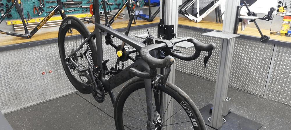 Ein schwarzes Rennrad ist in einer Werkstatt in einer Prüfstation eingespannt. Im Hintergrund hängen Werkzeuge an einer Metallwand, daneben stehen weitere Fahrradrahmen zur Begutachtung bereit.