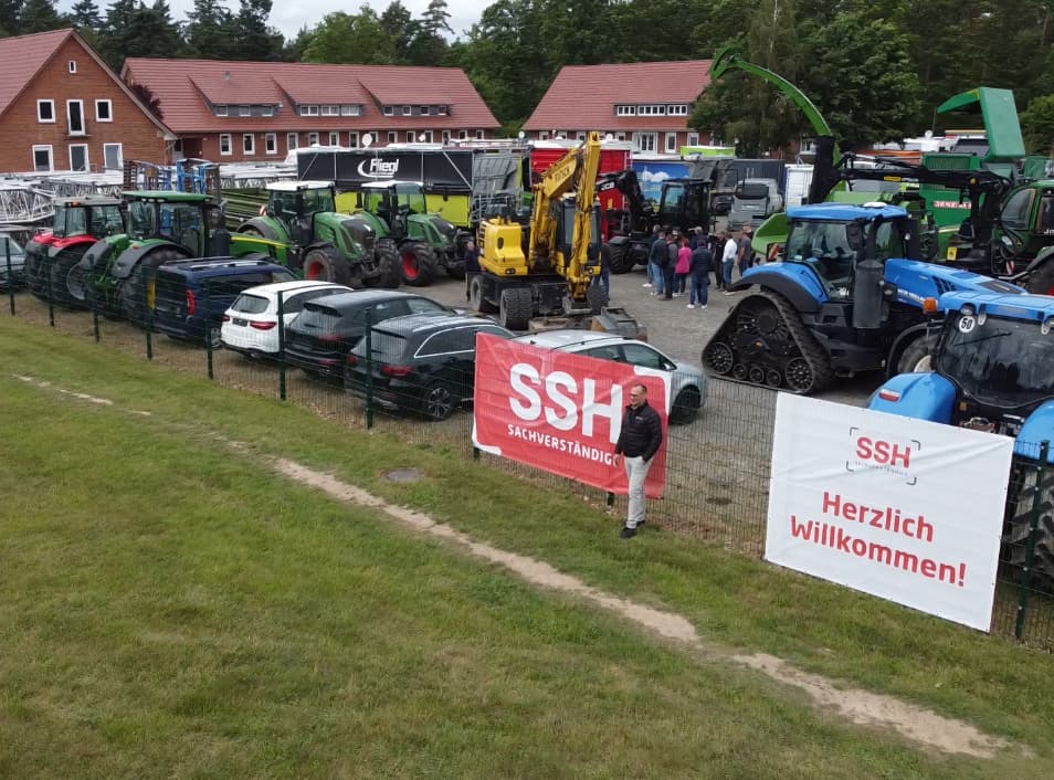  Übersicht über ein Ausstellungsgelände mit landwirtschaftlichen Maschinen und Fahrzeugen. Im Vordergrund hängen zwei Banner mit der Aufschrift „SSH Sachverständige“ und „Herzlich Willkommen!“ am Zaun, davor steht eine Person. Im Hintergrund sind Besucher, Traktoren und Baumaschinen zu sehen.