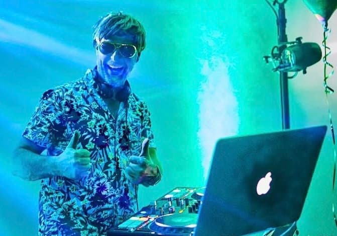  DJ Chicken Beats mit Sonnenbrille und Kopfhörern an einem DJ-Pult, zeigt Daumen hoch in blauer Lichtatmosphäre.