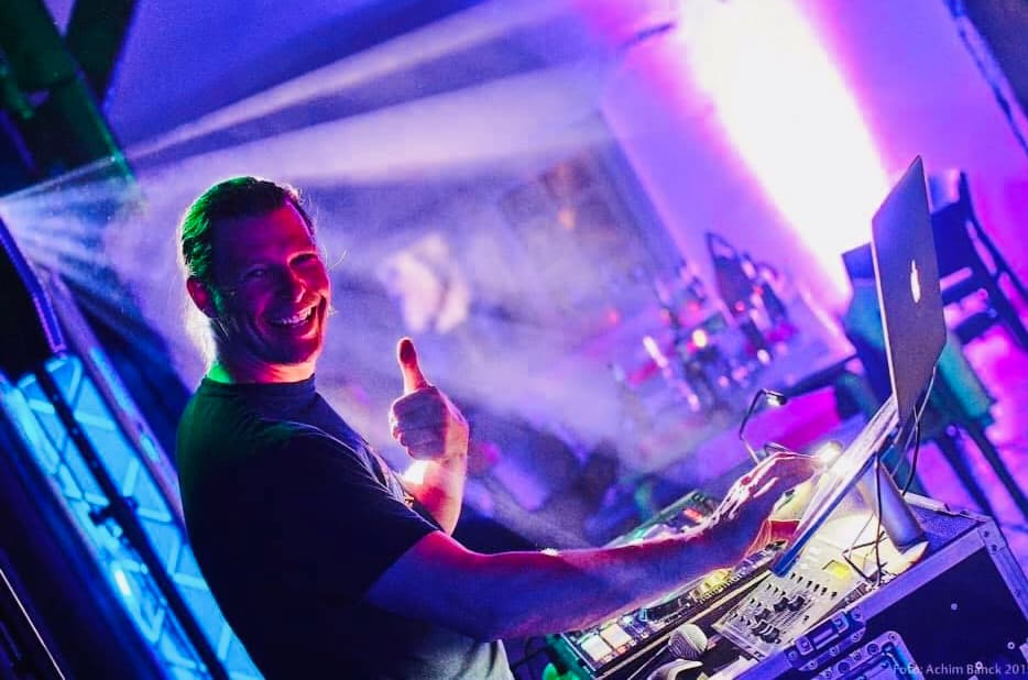  DJ Chicken Beats lächelt und zeigt Daumen hoch an seinem DJ-Pult, bunte Lichtstrahlen und Partystimmung im Hintergrund.