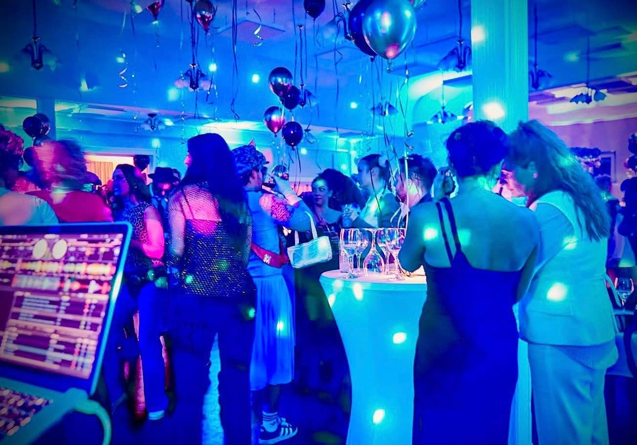  Menschen feiern ausgelassen auf einer Tanzfläche mit bunten Lichtreflexionen; DJ-Pult im Vordergrund sichtbar.