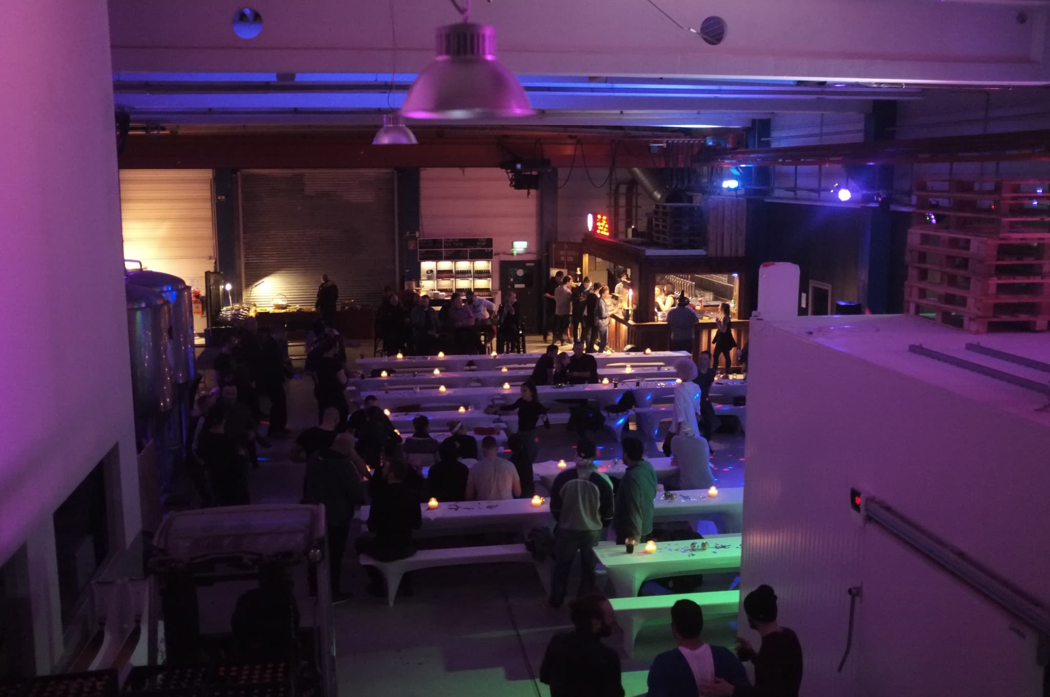 Blick von oben auf die Indoor-Eventfläche der Brauerei mit langen weißen Bänken, Kerzenlicht und bunt ausgeleuchteter Bar im Hintergrund.