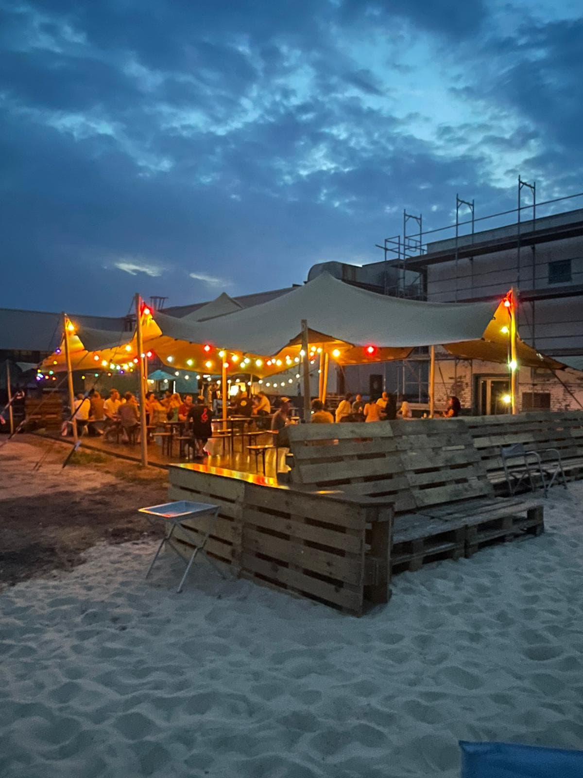 Abendstimmung im Outdoor-Bereich mit Sandboden, bunter Lichterkette und geselligem Zusammensitzen unter Zeltdächern – entspannte Beach-Atmosphäre.