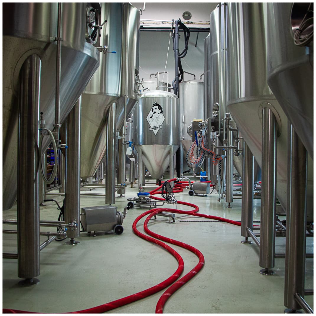  Blick in den Innenraum der Brauerei mit großen Edelstahltanks, roten Schläuchen am Boden – industrieller Charme trifft Handwerkskunst.