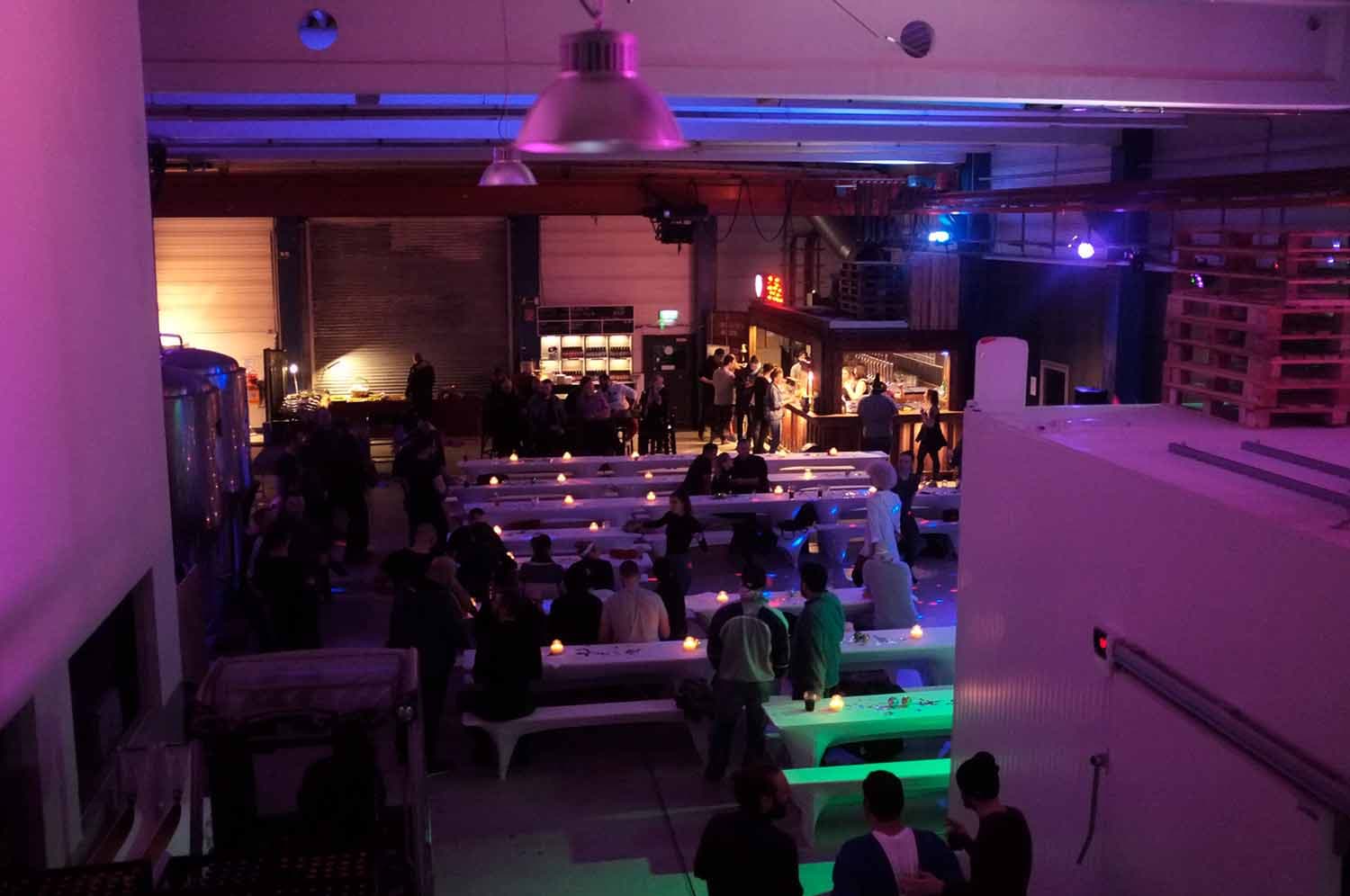 Blick von oben auf die Indoor-Eventfläche der Brauerei mit langen weißen Bänken, Kerzenlicht und bunt ausgeleuchteter Bar im Hintergrund.