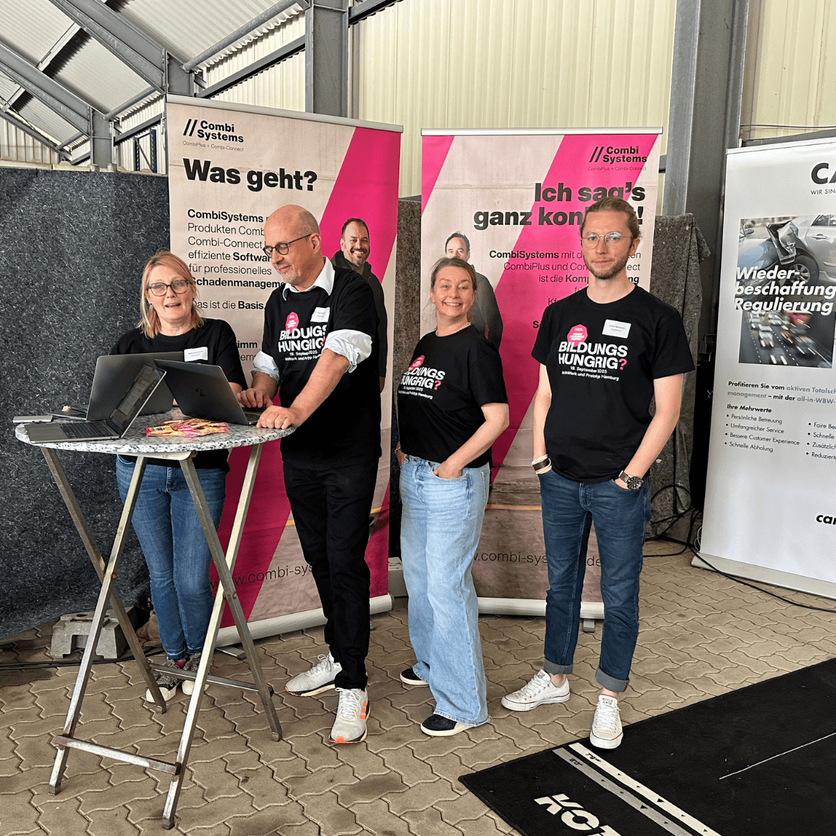 CombiSystems-Team beim SSH Praxistag für Kfz-Sachverständige in Leese. Das CombiTeam steht  am Stand von CombiSystems und lachen in die Kamera. Im Hintergrund sieht man die Chefs von CombiSystems auf den RollUps 